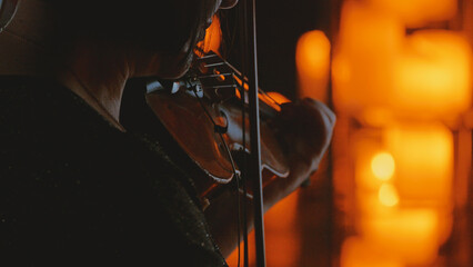 An intimate, close-up shot from behind a musician playing the violin. The scene is lit by warm, glowing candlelight, creating a dramatic silhouette and a moody, atmospheric bokeh background © VeNN