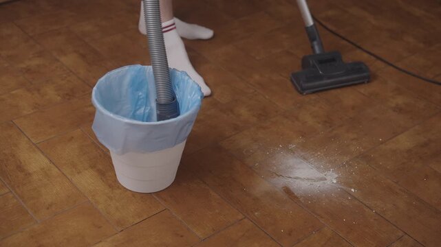 Person using vacuum cleaner to suck up dirt and debris from garbage bin, performing unique cleaning method ona traditional wooden floor, demonstrating innovative waste disposal, close-up, slow motion.
