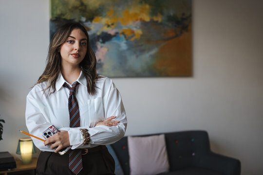 A young woman in a white shirt and tie stands with her arms crossed, holding a pencil and a phone.