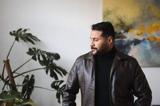 A man wearing a dark turtleneck and a brown leather jacket looks thoughtfully to the side, with a large plant and abstract art in the background.
