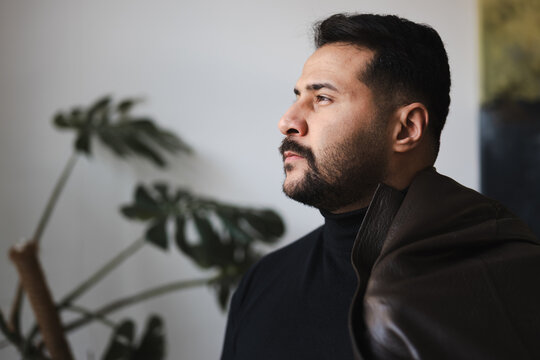 A thoughtful man with a beard wears a black turtleneck and a brown jacket draped over his shoulder, looking off into the distance.