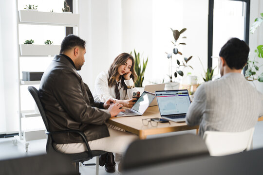 Three professionals collaborate around a table, reviewing data on laptops and a mobile device in a bright, modern office setting.