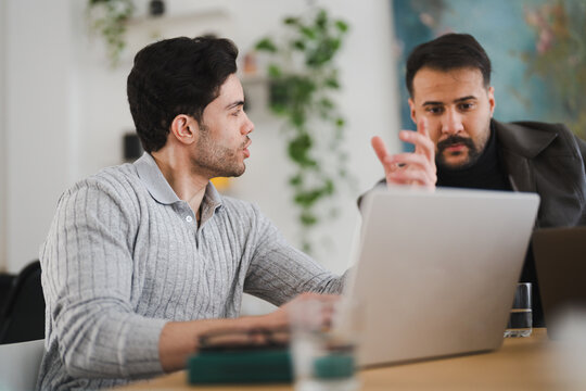 Two men collaborate on a project, one gesturing while discussing ideas with the other who is focused on a laptop screen.