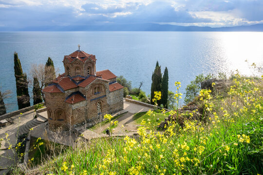 Iconic Saint John Kaneo church on a rocky outcrop above Lake Ohrid, framed by cypress trees and vibrant spring wildflowers, under a partly cloudy natural light.