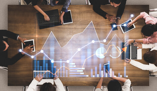 An overhead view of a business meeting featuring diverse professionals around a wooden table using laptops and tablets with data analysis graphics overlayed, symbolizing teamwork and strategy. Scalp