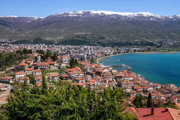 Naklejka premium A striking panorama of Ohrid's red-roofed buildings lining the lake's edge, backed by distant snow-capped peaks under a brilliant blue spring sky, framed by evergreen trees.