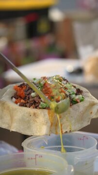 Cinematic 9:16 vertical slow motion shot of bright green salsa poured onto authentic al pastor tacos at a local Southern California taco vendor. Tight close-up angle emphasizes texture, freshness, and