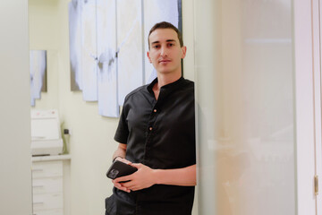 The dentist in the clinic stands leaning against the wall and holds a phone in his hands. A doctor in a black uniform. A handsome young doctor looks into the frame and smiles.