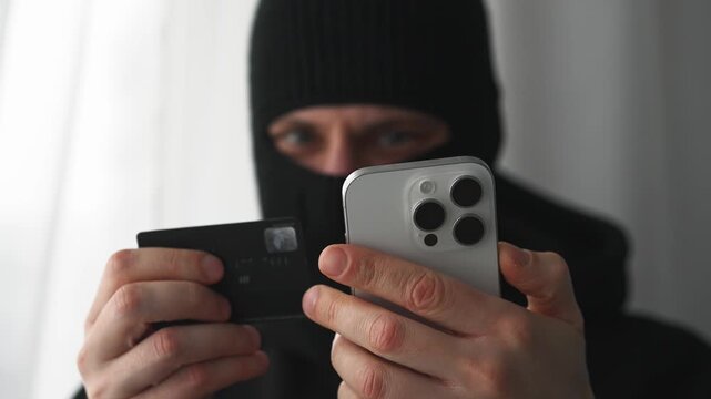 A scammer in a balaclava transfers money with someone else's phone.
Stealing money from a bank account.
A scammer in a black mask.