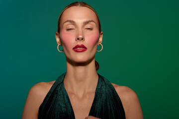 Portrait of a woman with closed eyes wearing dark green dress and gold hoop earrings on green background. Concept of elegance, fashion, beauty, and calm expression in studio lighting. © SHOTPRIME STUDIO