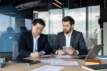 Diverse business professionals reviewing and signing document, sitting at meeting desk with laptop and tablet, working on partnership agreement in modern office