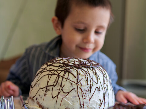 child looking down at a dome-shaped cake with white frosting and chocolate drizzle - A young boy aker admiring a finished homemade masterpiece in a domestic kitchen setting