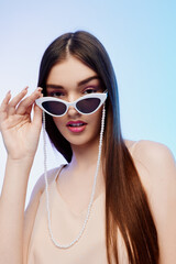 Fototapeta premium Young woman beauty fashion model with long straight hair wearing white cat eye sunglasses and a pearl chain. Studio portrait on a pastel gradient background. Concept for style and elegance.
