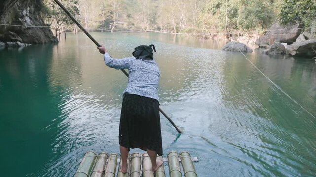 A woman poles a bamboo raft across a river in Hang Than Lim, Yen Minh, Ha Giang, Vietnam. She is transporting tourists the river in Ha Giang.