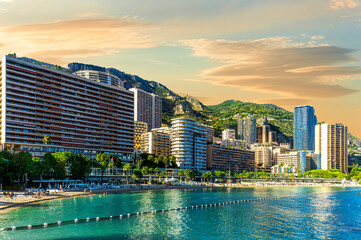 Obraz premium scenic cityscape of embarkment of Monte Carlo city in Monaco with blue sea water bay and amazing tall buildungs and houses along the coast. Urban mideterranean landscape with mountains and cloudy sky.