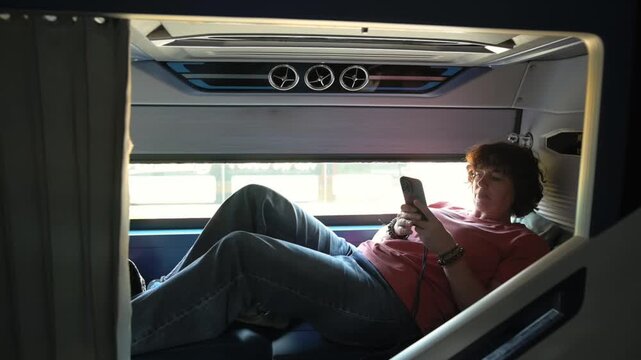 Sleeper Bus Passenger Relaxes During Travel. Traveler Resting And Checking Device. Young Traveler Stretching And Enjoying Sunlight. Passenger Relaxing In Sleeper With Sunlight And Listening