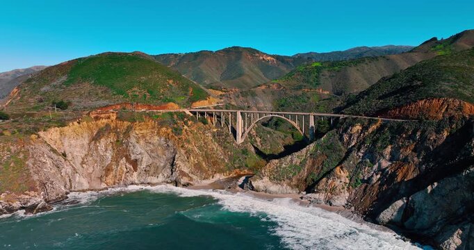 Bixby Creek Bridge aerial view with ocean waves and mountains. Historic concrete arch bridge spanning a canyon over a sandy beach in Big Sur