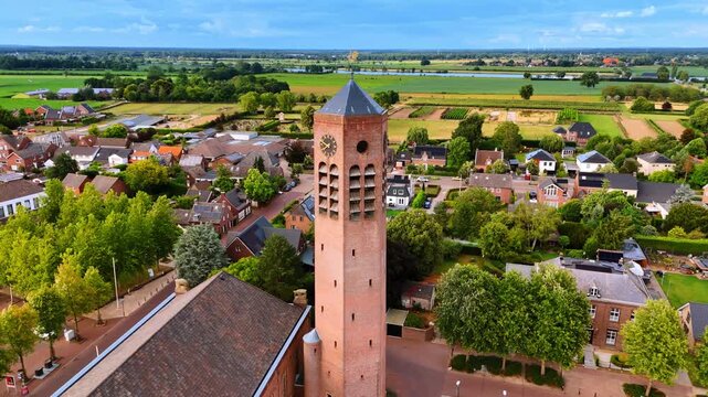 Aerial shot of traditional brick church tower over farmland. Detailed view of tall brick clock tower with residential houses and green fields in the background.