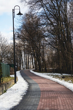 Winding paved walkway through a quiet park with snow along the edges, leafless trees, and vintage street lamps under a bright winter sky.