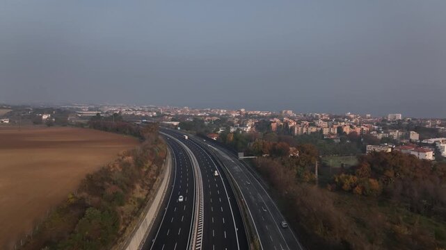 Early morning aerial drone flight over highways near Cingoli, Marche, Italy. Cars speed along the road with the town and Adriatic Sea visible in soft sunrise light.