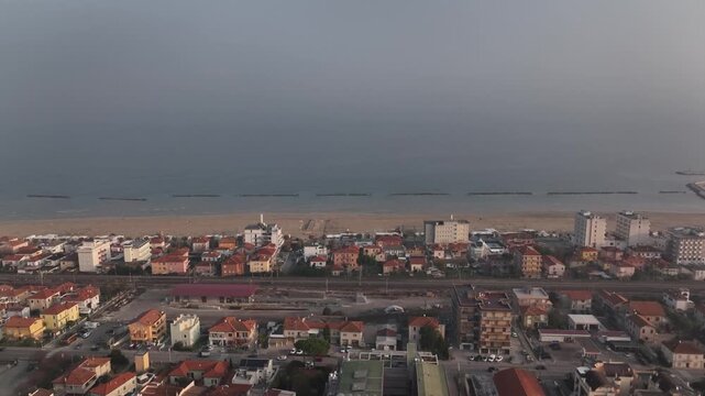 Early morning drone flight at sunrise over Cingoli, Marche, Italy. Light mist drifts above the town while the Adriatic Sea appears on the horizon in soft golden dawn light.