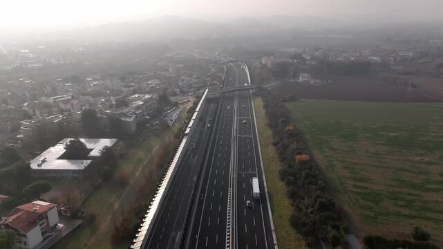 Early morning aerial drone flight over highways near Cingoli, Marche, Italy. Cars speed along the road with the town and Adriatic Sea visible in soft sunrise light.