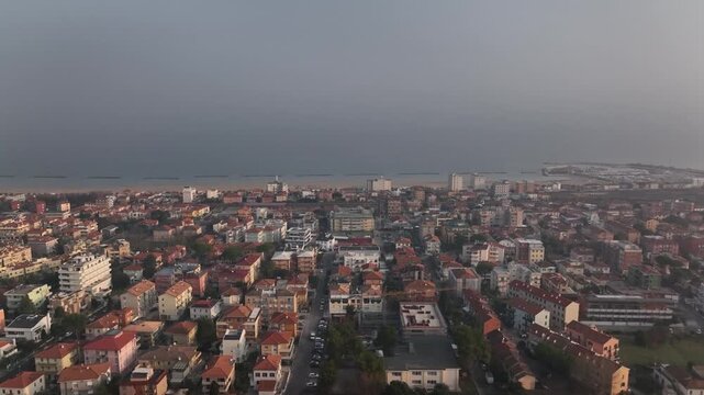 Early morning drone flight at sunrise over Cingoli, Marche, Italy. Light mist drifts above the town while the Adriatic Sea appears on the horizon in soft golden dawn light.