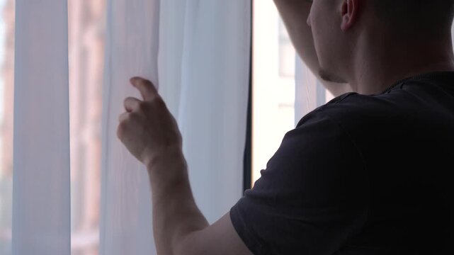 A man stands by the window, touching the sheer curtains.
Interior of a room.
White silk curtains in a cozy room.