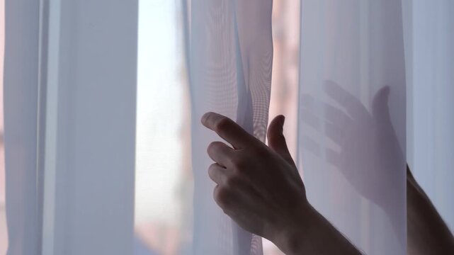 A man near a window.
The man touches the white, transparent tulle hanging on the window.