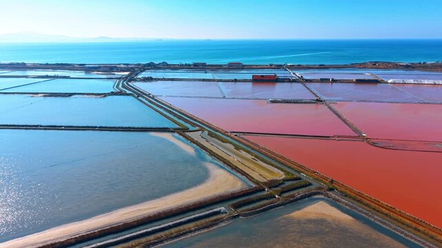 Wide aerial landscape of coastal salt production fields. High angle shot of pink and blue salt pans next to the sea with industrial buildings.