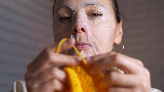 Woman knitting with cosmetic sheet mask on her face. Focused woman wearing a hydrating sheet mask on her face and knitting with yellow yarn, combining her skincare routine with a relaxing hobby