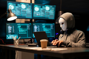 Masked male individual works on laptop at night, attempting to breach corporate systems. Anonymous hooded man appears to hack software company, representing cybercrime and digital security threats. © DC Studio