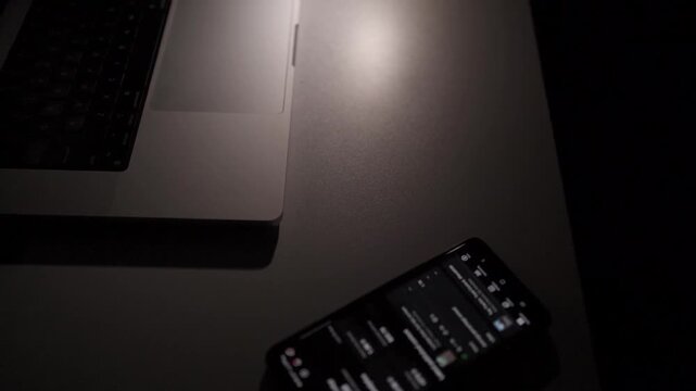 Laptop and smartphone with notification alert on desk in dark room. Concept of digital distraction, remote work pressure and always-on technology lifestyle.