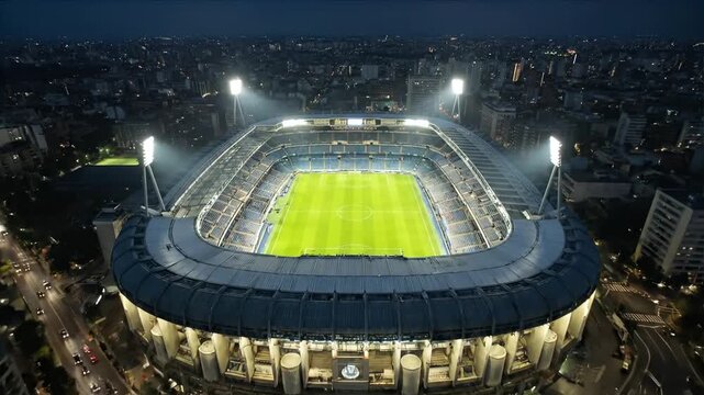 Aerial footage of a modern illuminated soccer stadium with green field at night, surrounded by a vast urban cityscape with numerous city lights