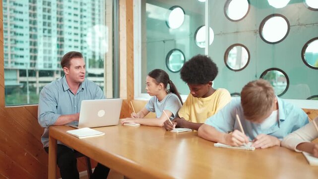 Smart teacher answer question while diverse student sitting and doing test. Multicultural children writing or taking notes while teacher using laptop analyze data and looking at paper. Edification