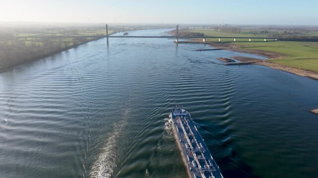 Aerial view of a wide river with two cargo ships leaving long wakes, surrounded by green fields, sparse trees, a distant bridge, and wind turbines across a calm rural landscape.