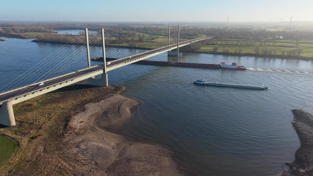 Aerial view of a cable stayed bridge spanning a wide river as two large vessels pass below, surrounded by green fields, forest patches, and distant wind turbines across a calm rural landscape.