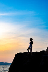 Beautiful young woman silhouette standing on stone at sunset or sunrise sky over sea
