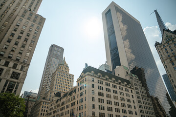 A view of modern and historic architecture in Midtown Manhattan under bright sunlight. Glass skyscrapers reflect the clouds while older stone buildings add contrast.