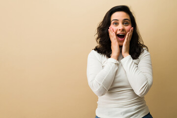 Hispanic woman showing surprise, mouth open and hands on face, experiencing unexpected shock and amazement
