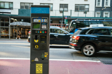 A modern parking meter stands on a busy New York City street with cars passing in the background. The machine accepts cards and coins for payment.