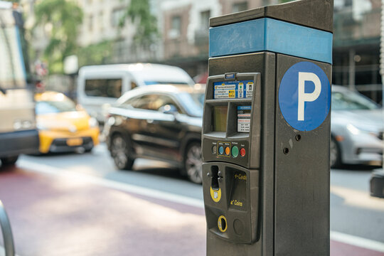 A close-up of a parking meter with a large "P" sign on a Manhattan street. Blurred traffic, including taxis and cars, moves in the background.