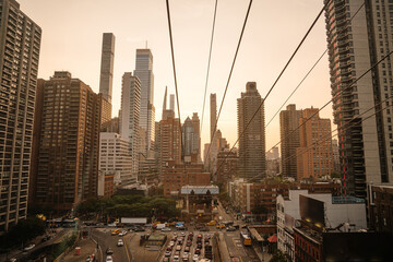 A dramatic view of the Manhattan skyline framed by tramway wires at sunset. Skyscrapers glow in warm light as traffic moves below in the heart of the city.