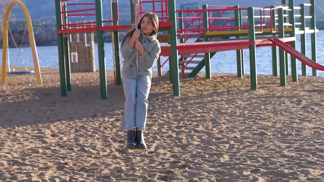 A young girl in cold weather clothing sitting on a rope hanging from a zipline, moving backwards. Playing on a playground on the beach of a lakeshore.