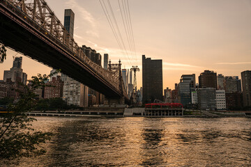 The Queensboro Bridge stretches over the East River at sunset with Manhattan skyscrapers in the background. The water reflects the golden sky, creating a peaceful urban scene.