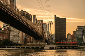 The Queensboro Bridge stretches across the East River at sunset with Manhattan's skyline glowing in the background. The Roosevelt Island Tramway and high-rise buildings create a striking urban