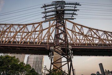 A detailed view of the Queensboro Bridge structure and the supporting tower for the Roosevelt Island Tramway. The strong steel framework and cables contrast with the modern skyscrapers and soft