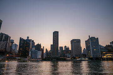 Tall buildings of Manhattan rise along the East River during dusk, with city lights beginning to glow. The calm water reflects the skyscrapers and the fading evening sky.
