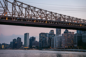 The steel structure of the Queensboro Bridge frames a view of Manhattan's skyline at twilight. The city's residential and office buildings are illuminated as day turns into night.