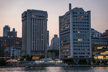 Two uniquely shaped concrete and glass office towers rise above the East River in Manhattan. The buildings are illuminated with warm lights as evening falls over the city.